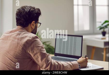 Back view of young business man working on a laptop with tables at office and writing. Stock Photo