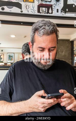 Concentrated bearded adult male in casual clothes looking at screen of mobile phone while standing in electronics shop and checking text messages Stock Photo