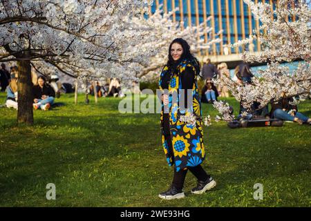 Portrait of a beautiful young brunette plus size woman with long hair, wearing a long yellow and blue vest, posing in a blooming garden Stock Photo