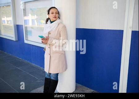 Young adult multi ethnic brunette, thoughtful woman in warm beige coat and denim, checking her smart mobile phone and looking into the distance while Stock Photo