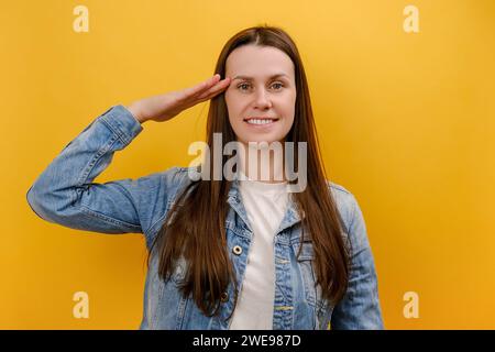 Portrait of cute young woman standing saluting with hand near head, looking obedient and attentive at camera, wearing denim jacket, posing isolated ov Stock Photo