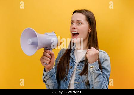 Portrait of angry young woman loudly screaming holding speaker in hand, strict boss yelling, raising fist, wearing denim jacket, isolated on yellow ba Stock Photo