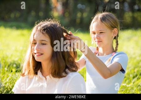 Two teenage girls spend time on green grass lawn in park, braid pigtails and tails for each other, enjoy summer and vacations Stock Photo