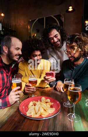 Group of friends in a bar having fun, drinking beer, using one phone watching social media. Stock Photo