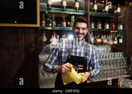 Young happy bartender cleaning the drinking glass. A handsome bartender polishes a glass of wine. The concept of service in coffee shop. Stock Photo