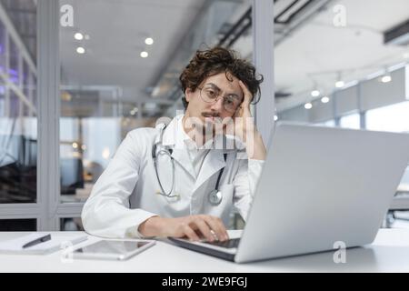 A man in a white medical coat with a stethoscope sits in front of a laptop computer in a medical office. Doctor thinking using computer Stock Photo