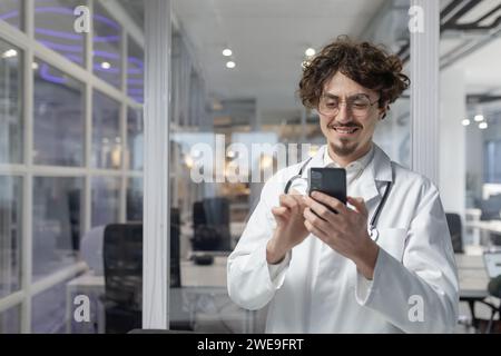 A man in a white lab coat and stethoscope holds a cell phone inside a medical office of a clinic. Stock Photo