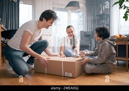 A boy and his parents are unpacking a large cardboard box at home together. Stock Photo