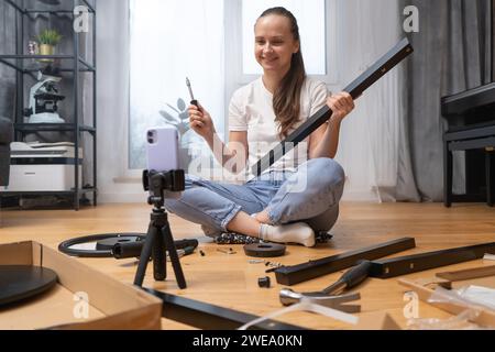 A vlogger girl records a video tutorial on furniture assembly on her phone. Stock Photo