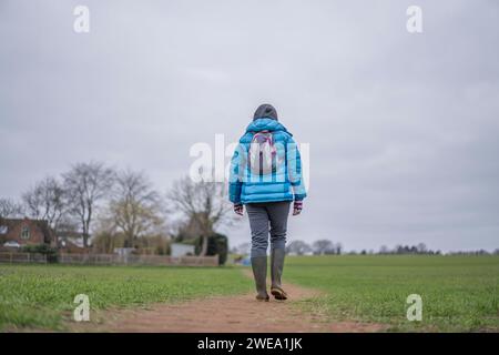 Rear view of a female walker strolling across a track in a farmers field with houses to the left side. Stock Photo