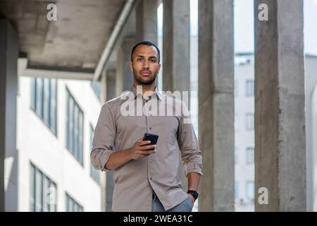 Serious and concentrated hispanic businessman looking at camera, man with phone in hands walking outside office building. Stock Photo