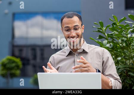 Close up of young student using laptop for video call, joyful man smiling talking to fellow students sitting on bench outside university campus Stock Photo