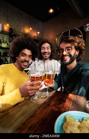 Selfie of three male friends toasting happily in a bar, looking at camera cheerfully. Stock Photo