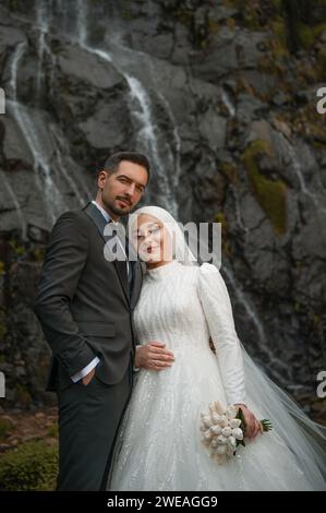 Happy bride and groom standing in front of a waterfall looking at camera smiling and posing Stock Photo