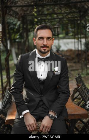 Handsome groom in black suit sitting on park table looking at the camera and posing in the park Stock Photo