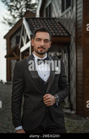 Handsome young groom in black suit standing, looking at the camera, posing outdoors Stock Photo