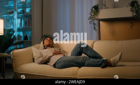 Tired African American woman in evening night living room lying on cozy sofa falling down on comfortable soft couch relaxed tranquil calm girl dream Stock Photo