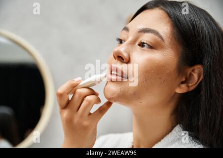 young and brunette asian woman with acne applying lip balm and looking at mirror in bathroom Stock Photo