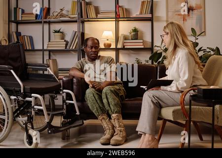 Black war veteran with disability sitting on couch and talking to psychologist listening to him Stock Photo