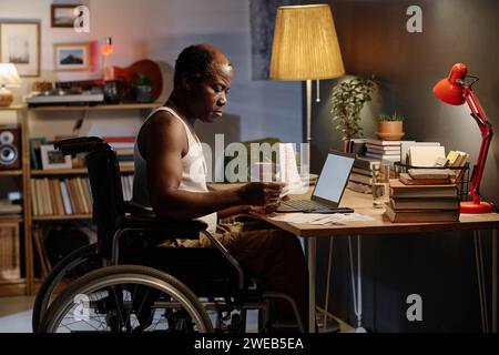 Mature african american man in wheelchair sitting at desk in his bedroom and looking through bills Stock Photo