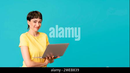 Smiling woman in a yellow t-shirt using laptop, pc computer on blue background with copy space. Work studying, virtual training, e-learning, watching Stock Photo