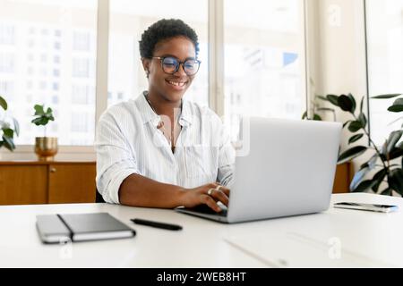 Focused businesswoman using laptop computer in contemporary coworking space, freelance programmer in glasses coding sitting at the office desk, creativity designer or architect typing Stock Photo