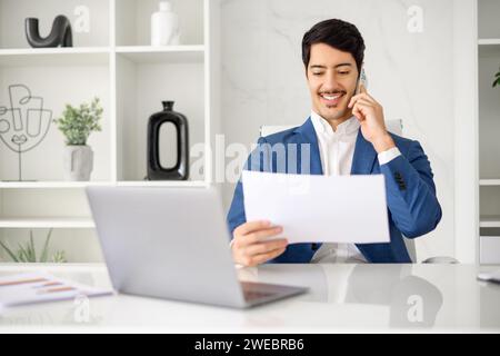 Hispanic businessman attentively examines a document while on a phone call, capturing the multitasking nature of the business environment, male office employee talking while looking through contract Stock Photo