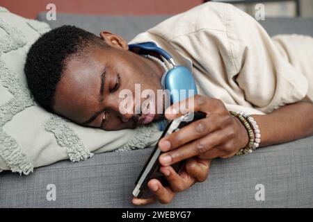 Restful African American guy relaxing on couch in front of camera and looking at screen of smartphone on gamepad during video game Stock Photo