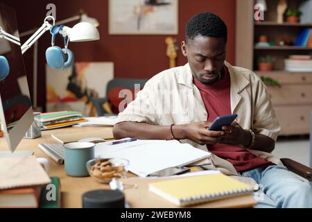 Young male freelancer looking at smartphone screen while sitting by workplace with papers and notepads and organizing work Stock Photo