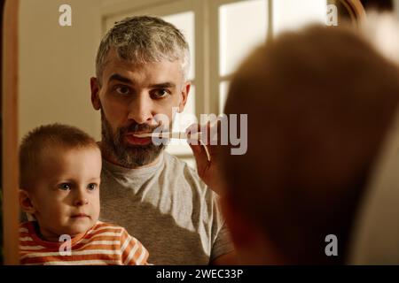 Mature bearded father brushing teeth with toothbrush in front of mirror during morning hygiene while his cute baby son looking at him Stock Photo