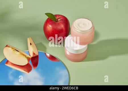 Round mirror with two apple slices displayed on. Two pink jar containing cream inside stacked on each other. Natural beauty blank label for branding m Stock Photo