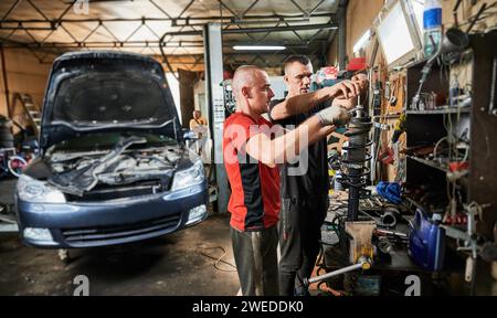 Side view of two men working in garage, auto, servicing center. Professional workers, repairmen, mechanics fixing, repairing car, holding equipment. Concept of manual labor. Stock Photo