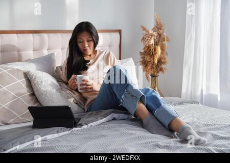 Attractive woman in casual clothes holding cup working on digital tablet sitting on bed Stock Photo