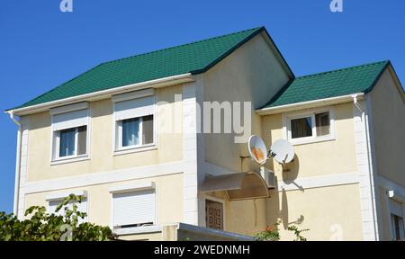 A modern detached house with a green metal roof, stucco, plastered painted walls, windows with closed plastic rolling shutters, and two satellite dish Stock Photo