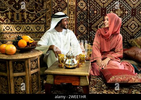 Young couple from Emirati spending time in an arabian traditional cafe. Man and woman wearing kandura and abaya from Dubai conversating together. Stock Photo