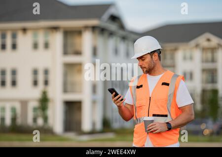Engineer builder drinking take away coffee using phone on break. Builder at construction site. Buider with helmet on construction outdoor. Worker at Stock Photo