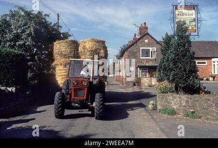 1990s UK farmer harvesting, bring bails of hay in from the field driving tractor past The Plough Inn,  Wistanstow, Shropshire, England  circa 1991. Stock Photo