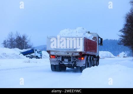 Tipper truck transporting a load of cleared snow away from town to snow dump in winter snowfall, rear view. Another truck unloading. Stock Photo