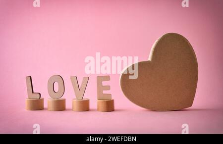 Love background with cutouts of letters forming word love on wooden blocks and brown heart-shaped gift box on pink background. Front view. Stock Photo