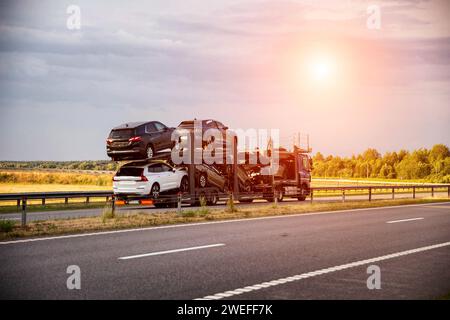 An auto transporter semi-trailer truck transports broken damaged cars from Europe in the summer against a sunset background. Car import and export con Stock Photo