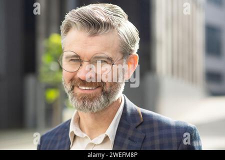 Experienced senior man with a warm smile standing confidently outside an office, dressed in a professional business suit. Stock Photo