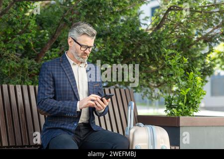 Mature man in a business suit using a phone while sitting on a bench outside an office with a suitcase, portraying a professional on the move. Stock Photo