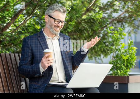 Experienced senior banker in a stylish suit enjoys a coffee break while working on a laptop outside the office building. Stock Photo