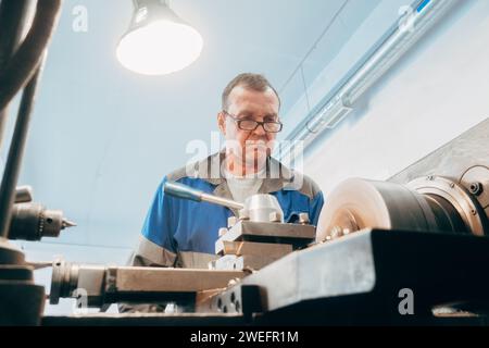 Elderly turner, 50-55 years old, works at lathe in turning shop. Front view. Authentic workflow. Stock Photo
