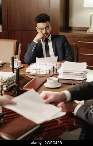 Young attorney reading document sitting at desk in office boardroom, his coworkers exchanging papers Stock Photo