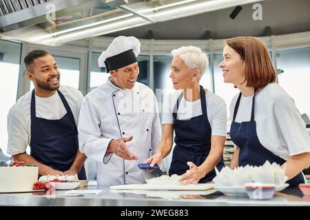 mature beautiful woman in apron working with dough and looking at cheerful interracial chefs Stock Photo