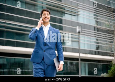 A proud Hispanic professional in a blue suit engages in a conversation on his smartphone, carrying a laptop, representing the importance of communication and mobility in the fast-paced business world Stock Photo