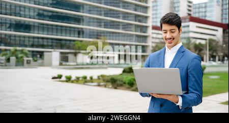 A young Hispanic professional, elegantly dressed in a blue suit, works on his laptop against a backdrop of urban architecture, epitomizing mobile business operations. Business and technology concept Stock Photo