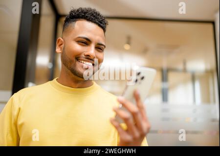 Cheerful Indian male employee, freelancer using cellphone and smiling, reading good news message, enjoying mobile application. Man looking at phone screen with toothy smile, standing indoors Stock Photo
