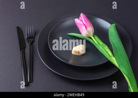 Pink tulip flower heart shaped candy on black plate on black background. Valentines day, womens day concept. Top view. Stock Photo